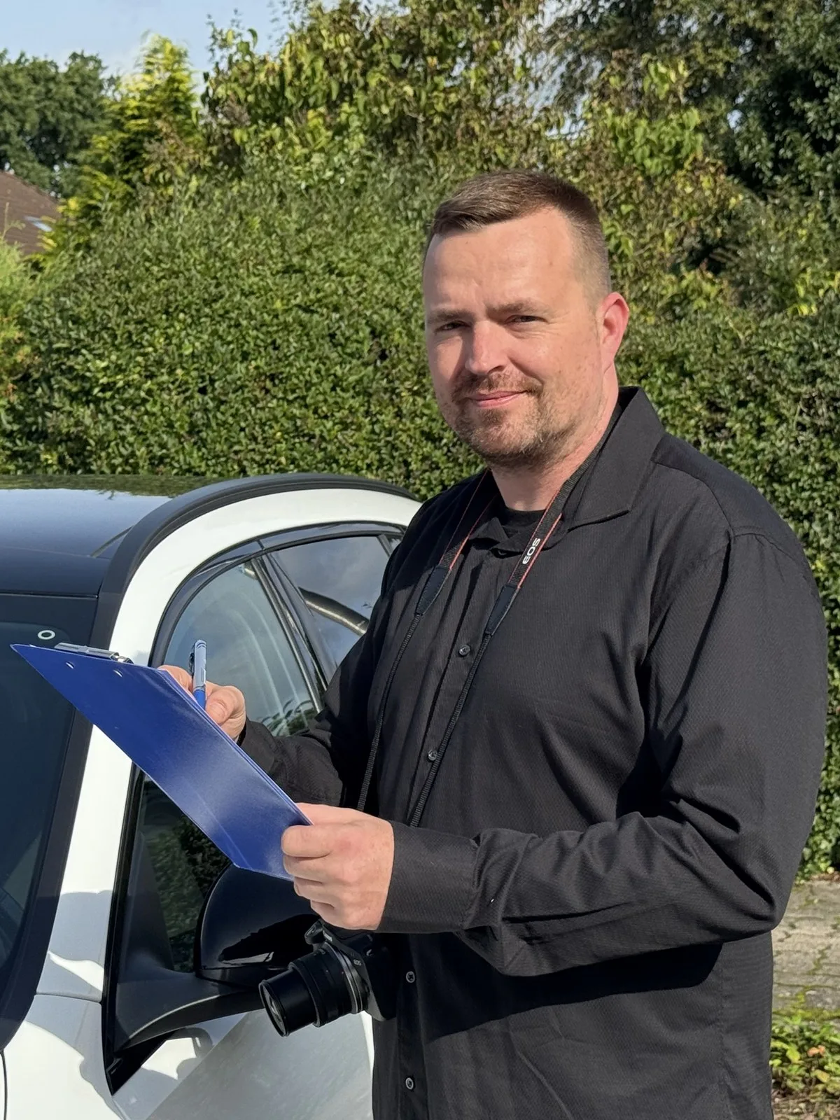 KFZ-Sachverständiger Marco Birkholz mit Klemmbrett und Kamera an einem weißen Auto